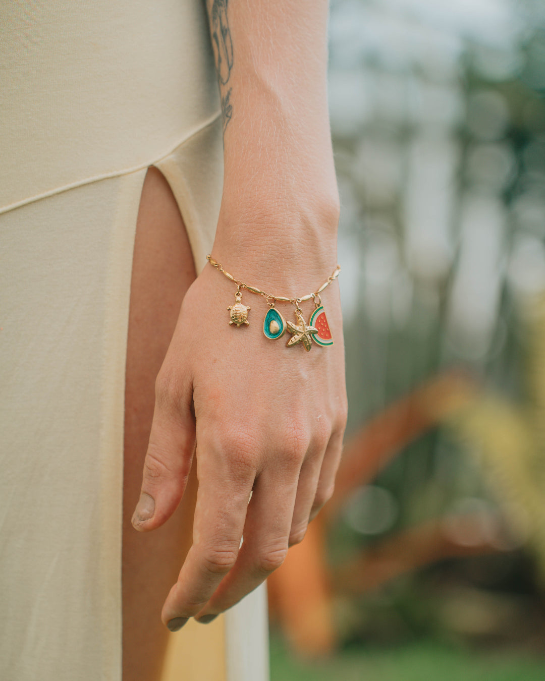 Hand wearing a gold bracelet with charms against a blurred outdoor background